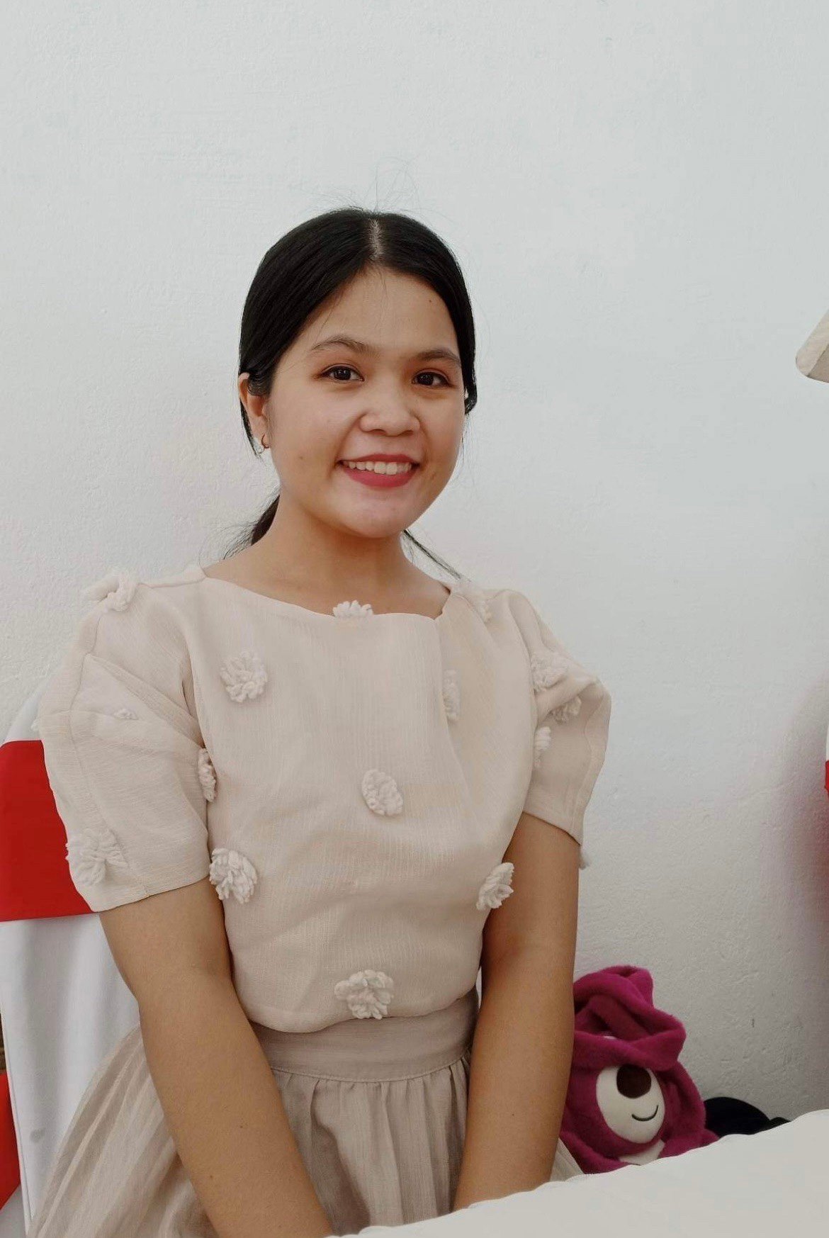 Smiling woman in beige textured dress sitting indoors with plush toy nearby