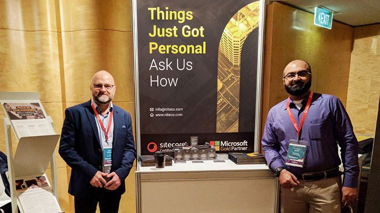 Men at a tech event booth with "Things Just Got Personal" banner and Microsoft partner logos