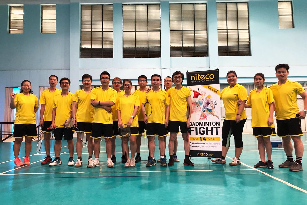 Badminton team in yellow uniforms posing by Niteco Badminton Fight event banner